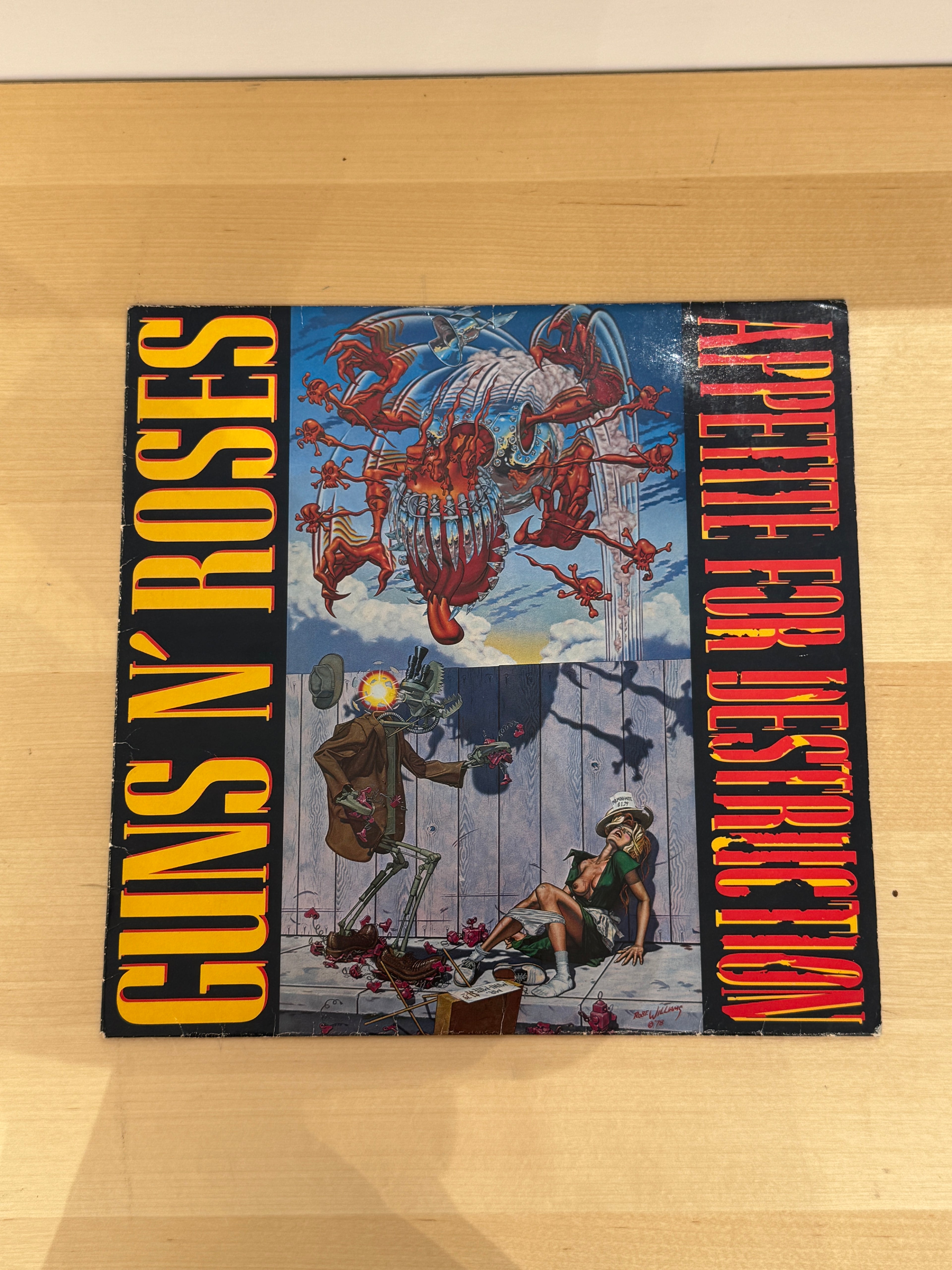 Guns N' Roses – Appetite for Destruction, copertina fronte prima stampa europea 1987: artwork Robert Williams con robot e scena violenta su sfondo cielo blu, titolo verticale giallo-arancio a sinistra, titolo verticale rosso-rosa a destra, sfondo nero