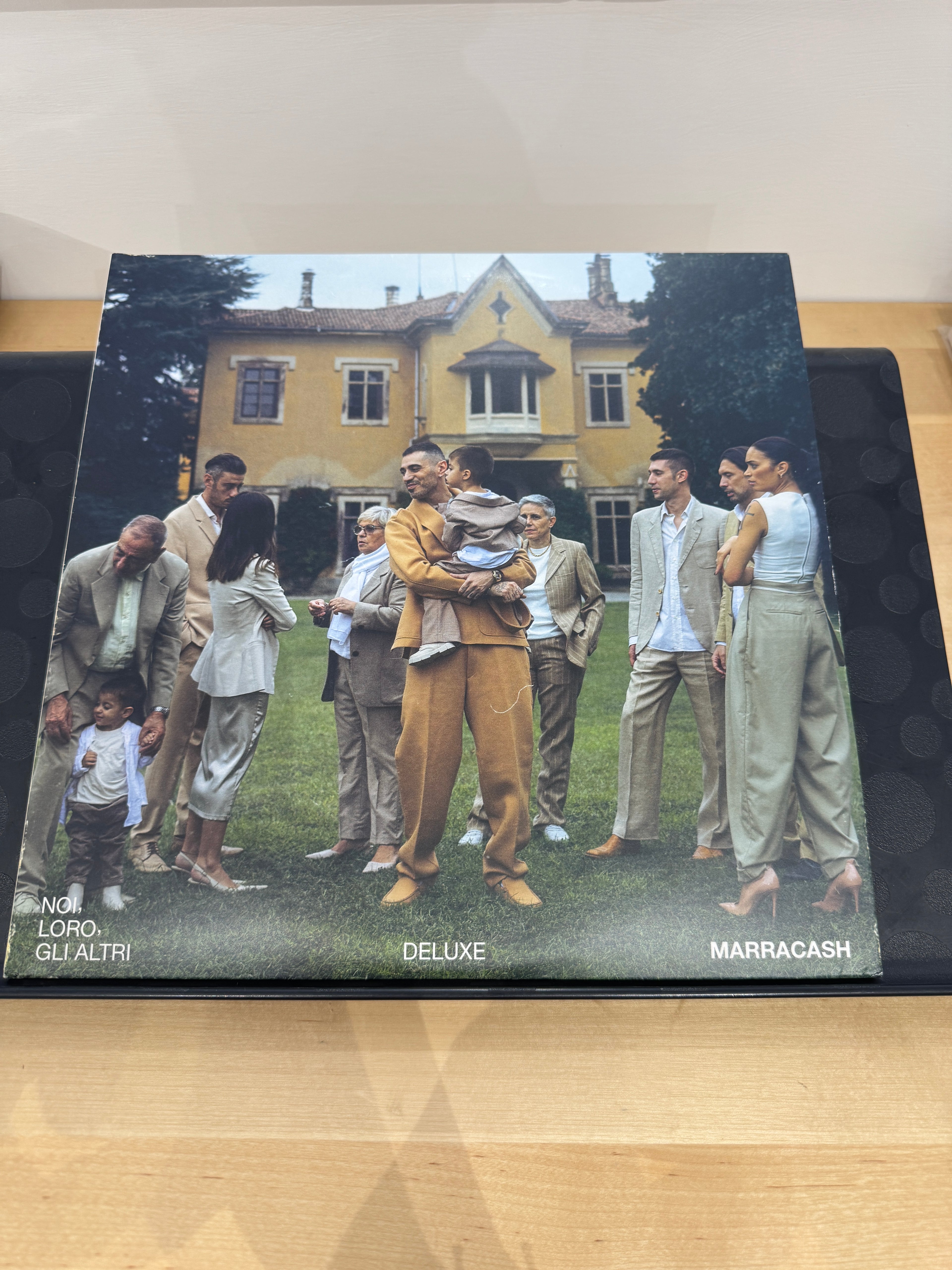 Copertina Marracash Noi Loro Gli Altri Deluxe Edition - foto di gruppo con Marracash al centro che tiene in braccio un bambino davanti a una villa, scritta Deluxe in basso